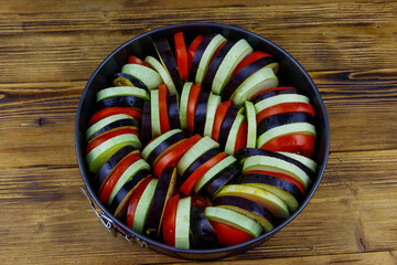 Preparing of ratatouille. Raw vegetables for ratatouille in round baking dish on wooden table....