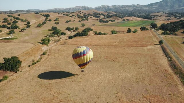 Aerial 4K overhead view of descending hot air balloon full of happy excited people tourists, visiting sunny summer California, Solvang area USA. Scenic countryside view with green mountains background
