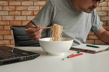 Thai male worker busy working with laptop, use chopsticks to hastily eat instant noodles during...