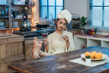 carefree asian female with towel on head and legs on dining table is reading social media feeds on...