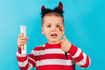 One little toddler boy with horns on his head and with toothpaste and a brush in his hands is isolated on a blue background. Health, children's hygiene, holidays concept.