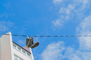 Two pairs of sneakers casted upstairs, hang with soles down on twisted black cable stretched at height on background of blue suburban sky. Upper floors of residential building are visible on the left.