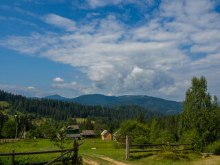 Fototapeta na wymiar Carpathian landscape with cloudy sky. A wooden house on a green meadow in mountains near old forest. Lifestyle in the Carpathian village. Ecology protection concept. Explore the beauty of the world.