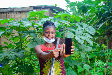 A female african farmer with nose mask and a farming hoe on her shoulder happily shows and points to the smart phone she is holding in a farm or garden - obrazy, fototapety, plakaty