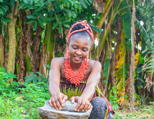 A pretty African lady or woman with beads on her head is grinding something with local grinding stone in a banana farm - obrazy, fototapety, plakaty