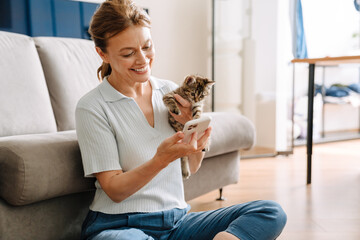 Senior woman holding kitten and using cellphone while sitting on floor © Drobot Dean