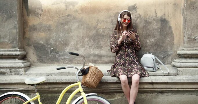Beutiful girl is enjoying the music she is listening to. Also she is dansing and pointing up. Girl is enjoying the moment. Yellow bike, red glasses, white headphones, white bagpack