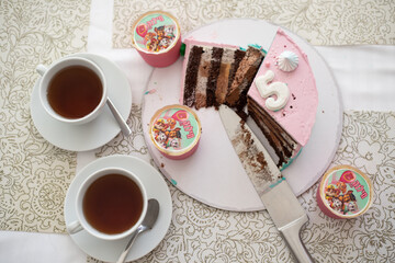Piece chocolate cake with many layers of cream decorate with pink mastic. Cut piece. Big knife and white two cups of tea. Top view. Birthday cake 5 years
