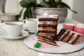 Piece chocolate cake with many layers of cream decorate with pink mastic. Cut piece. Two white cups of tea, plate with cake and pot with flower on table. Side view. Cake on holiday.