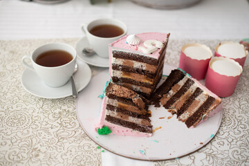 Pieces chocolate cakes with many layers of cream decorated with pink mastic. Cutted pieces cake on white plate and two white cups of tea. Top view. Birthday cake 5 years.