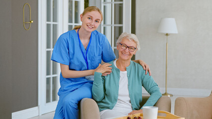 Smiling young blonde nurse in uniform takes care of senior woman sitting in comfortable armchair in light room