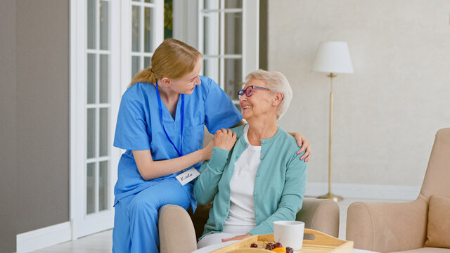 Smiling blonde nurse in uniform takes care of senior woman sitting in comfortable armchair