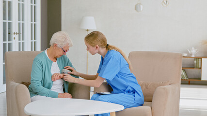 Young nurse in blue uniform puts digital blood pressure monitor on senior woman hand in light office