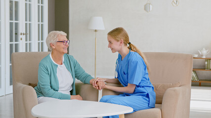 Positive doctor joins hands cheering up mature grey haired lady sitting in armchairs in stylish office - 459842937