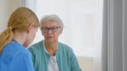 Senior lady patient with glasses talks to experienced young caretaker sitting near window in light room