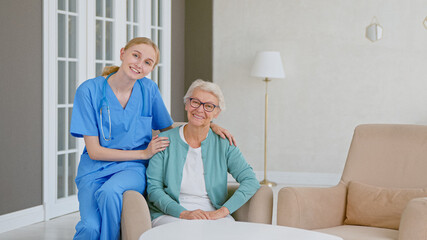 Young blonde nurse in uniform takes care of senior woman in light room at assisted living - 459843761
