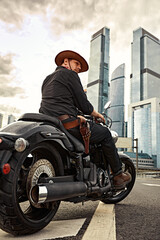 Fototapeta premium Portrait of stylish male dressed in a black leather jacket and bandana with gun sitting on his custom-made retro motorcycle, looking away, buildings of the big city on background.
