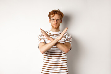 Serious young man with ginger hair frowning, showing stop gesture, making cross to prohibit...