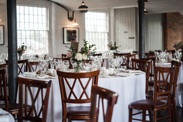 Romantic wedding breakfast room set up for guests and the bride and groom.  Wooden chairs, tables, glasses flowers and industrial setting.