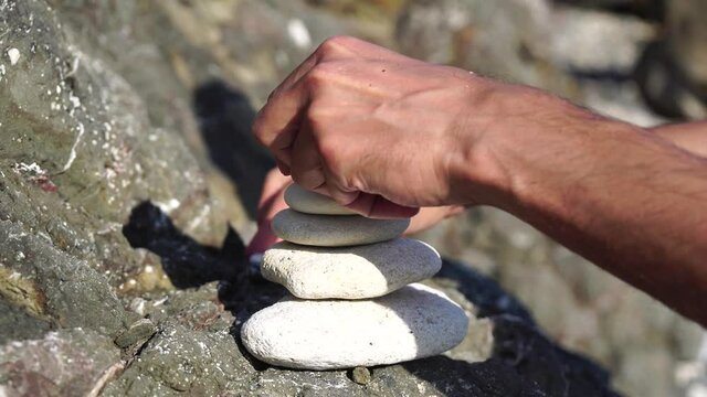 Man bilds stones pyramid on the seashore on a sunny day on the blue sea background. Happy holidays. Pebble beach, calm sea, travel destination. Concept of happy vacation on the sea, meditation, spa
