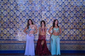 Three middle-aged Hispanic women in beautifully colored and beaded dresses for belly dancing, posing with a wall of beautiful tiles in the background. Belly dance concept.