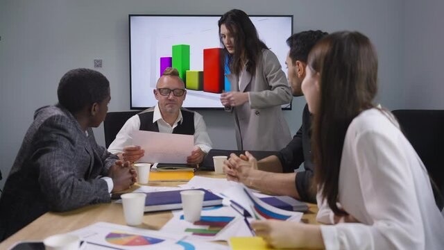 Male CEO in eyeglasses talking with female manager discussing business profitability report in office with multiethnic colleagues sitting at table. Confident intelligent people planning strategy