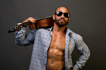 Cool muscular Latin American in sunglasses and a shirt holds an acoustic guitar on his shoulder and shows a peace sign on a gray background