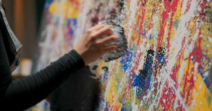 Art student spends time at university in painting studio, completing her new project, a modern, abstract work of oil paint, stamping off bubble wrap soiled with black paint she picks up from bucket
