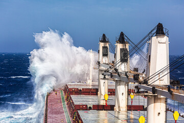 A big wave hits the cargo ship with cargo cranes in the Bow and rises several meters above it. 