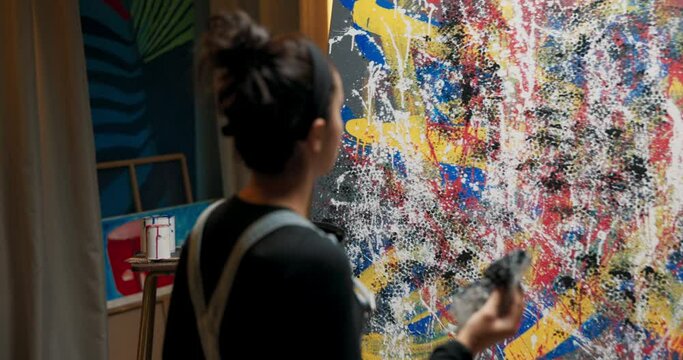 Women with artistic soul spend night in painting studio amidst chaos of paints brushes stands at easel with big abstract painting dips bubble wrap into large bucket of black paint and stamps on canvas