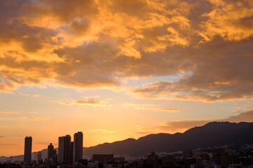 Fototapeta premium 都会の夕暮れ。六甲山に沈む夕陽とオレンジ色に染まる空と雲。高層ビルがシルエットで浮かぶ。