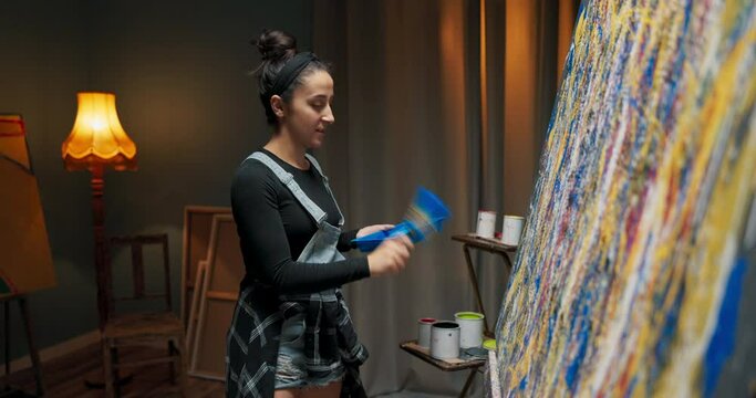 Dark studio lit by lamp at night woman artist painter stands by canvas with abstract painting in hand she holds container with blue paint and brush applies with quick movement