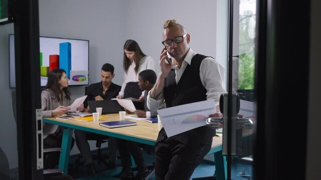 Portrait of serious confident man talking on phone analyzing graph with blurred colleagues at background in meeting room. Professional Caucasian expert with multiethnic coworkers in office indoors