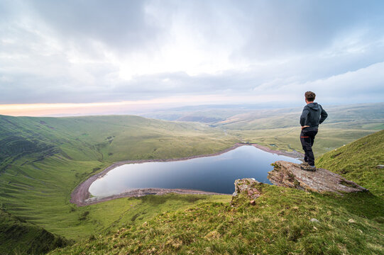 Man standing on rocks overlooking Llyn y Fan Fach lake. Brecon Beacons National Park. Black Mountain, Carmarthenshire, South Wales, the United Kingdom. Hiking in the UK concept.