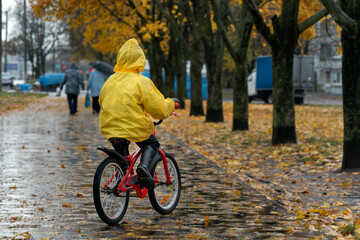 Back view on the child in yellow raincoat. Boy rides bicycle in an autumn park in the rain.