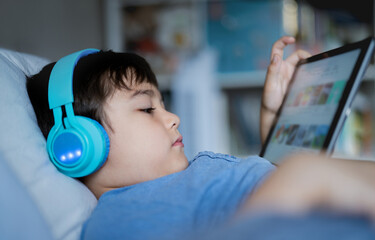 Close up face young boy wear wireless headphone listening music while sitting in bed,Cute kid playing game on tablet,Positive child in blue pajamas relaxing in bed room in morning before go to school