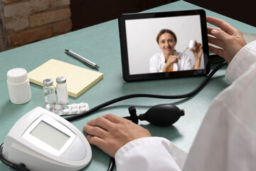 Video call between two doctor who discussing various ways to lower blood pressure.Tablet pc,blood pressure monitor,pills,bottles on the blue table and doctor sitting at the table