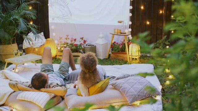 Blonde woman and husband watch movie on large screen hung on fence while lie on rest place with plaid and pillows in cottage yard backside view
