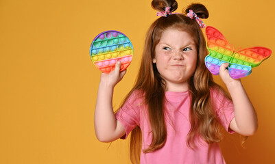 Frolic grimacing red-haired kid girl in pink shirt holds two sensory rainbow color toys, round and butterfly share pop it and looks aside at round one