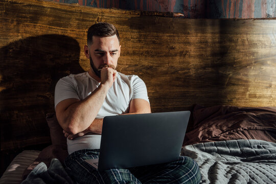 Thoughtful Man Looking At Laptop On Bed