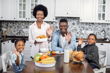 Portrait of young parents with two cute daughters smiling and waving hands on camera while sitting together on bright kitchen. African american family having breakfast at cozy home.
