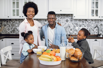 Front view of young african parents with two pretty daughters sitting at kitchen table with fresh fruits, juice and cookies. Breakfast time of happy family. Domestic lifestyles.