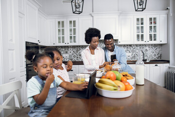 Two african sisters sitting on chairs and watching cartoons on digital tablet while their young parents using smartphone on background. Family with gadgets on kitchen. Modern lifestyles.