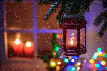 Close up of a Christmas lantern hanging from a pine branch on a snowing night. A window lit by...