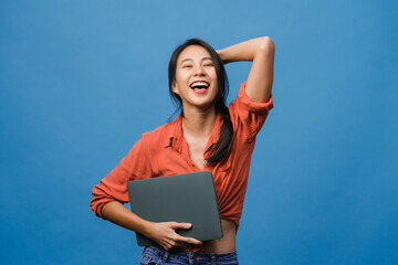 Obraz premium Surprised young Asia lady hold laptop with positive expression, smile broadly, dressed in casual clothing and looking at camera on blue background. Happy adorable glad woman rejoices success.