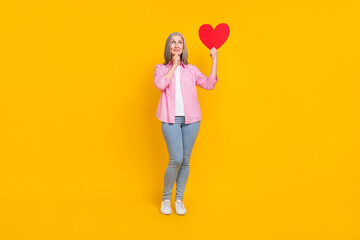 Full body photo of mature woman happy positive smile think hold paper heart love isolated over yellow color background