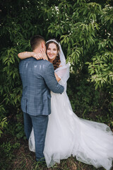 A cute, beautiful bride in a white dress and a long veil hugs the stylish groom behind his back in nature with green leaves. Wedding photography.