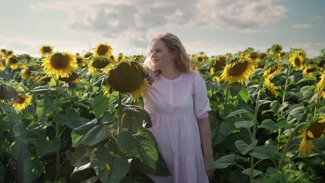 a young pretty blonde caucasian girl with curly hair in a light dress at a photo shoot in a sunflower field on a sunny day smiles, posing on the camera