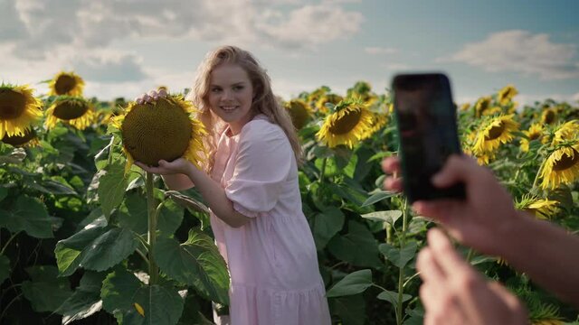 a young pretty blonde caucasian girl with curly hair in a light dress at a photo shoot in a sunflower field on a sunny day smiles, posing on the phone camera to a cute guy