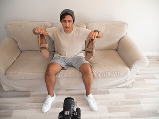 Hispanic young man creating clothing content for social networks with a camera on a tripod, sitting on a piece of furniture